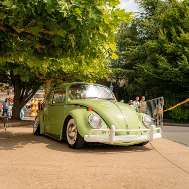 Käfer in grün parkt unter einem Baum, im Hintergrund Menschen und eine Straße.